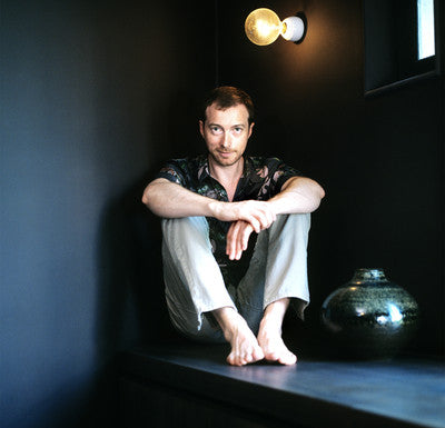 Christophe Delcourt sitting on a ledge in a dimly lit room with a light fixture and vase.
