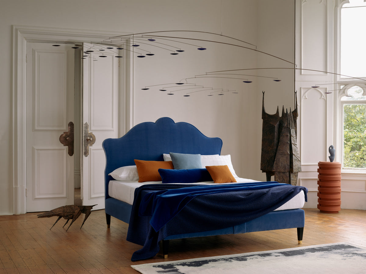 Blue upholstered Lenoir headboard in a stylish bedroom with large mirror and decorative elements.