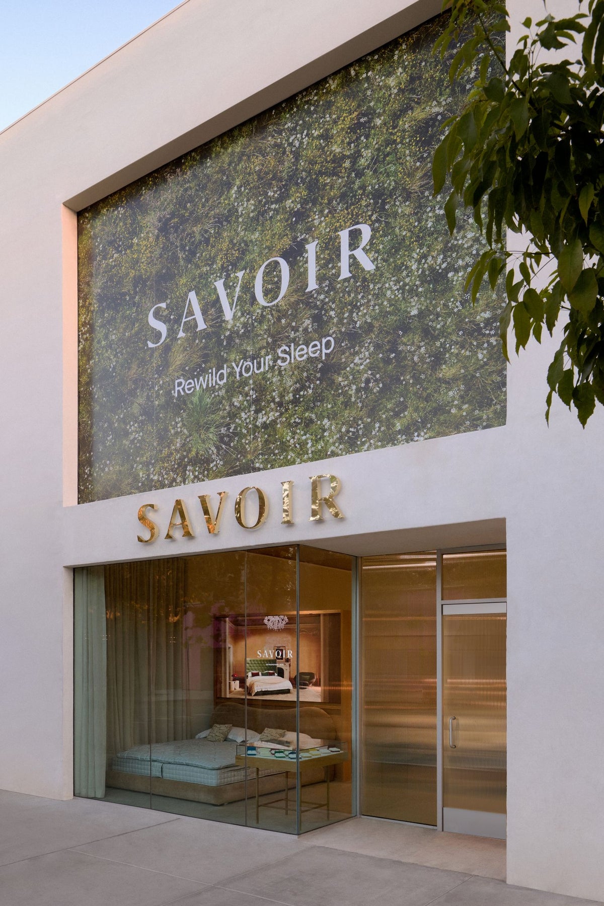 Savoir Los Angeles showroom exterior with a large green textured wall panel reading 'SAVOIR Rewild Your Sleep' and gold 'SAVOIR' lettering above glass doors and display windows showing beds inside.