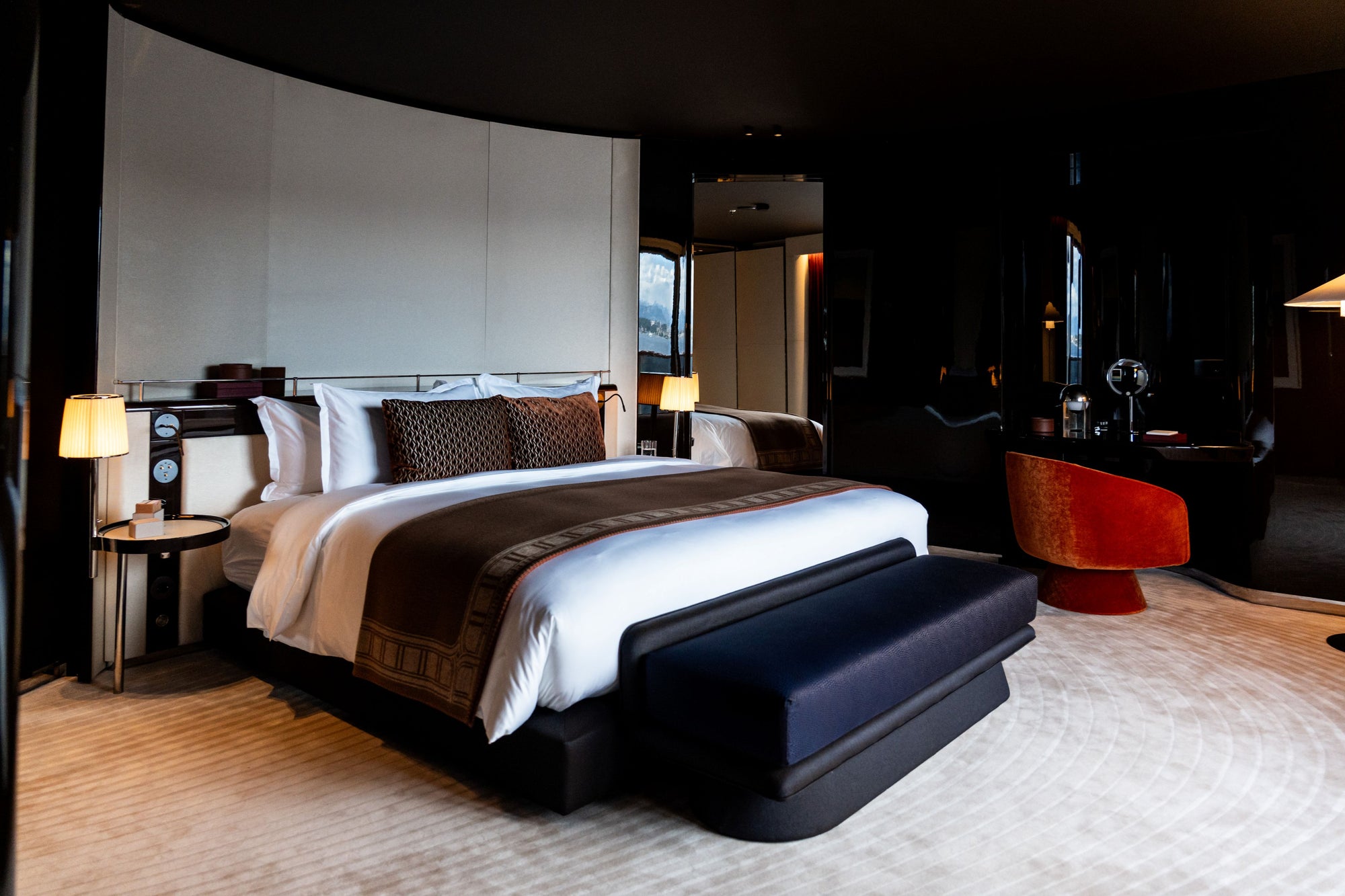 A luxury modern bedroom in a yacht featuring a large bed dressed in white linens with dark brown patterned accents cushions, set against with bedside tables and lamps. A dark upholstered bench sits at the foot of the bed, and a vibrant orange-red accent chair is visible in the background.