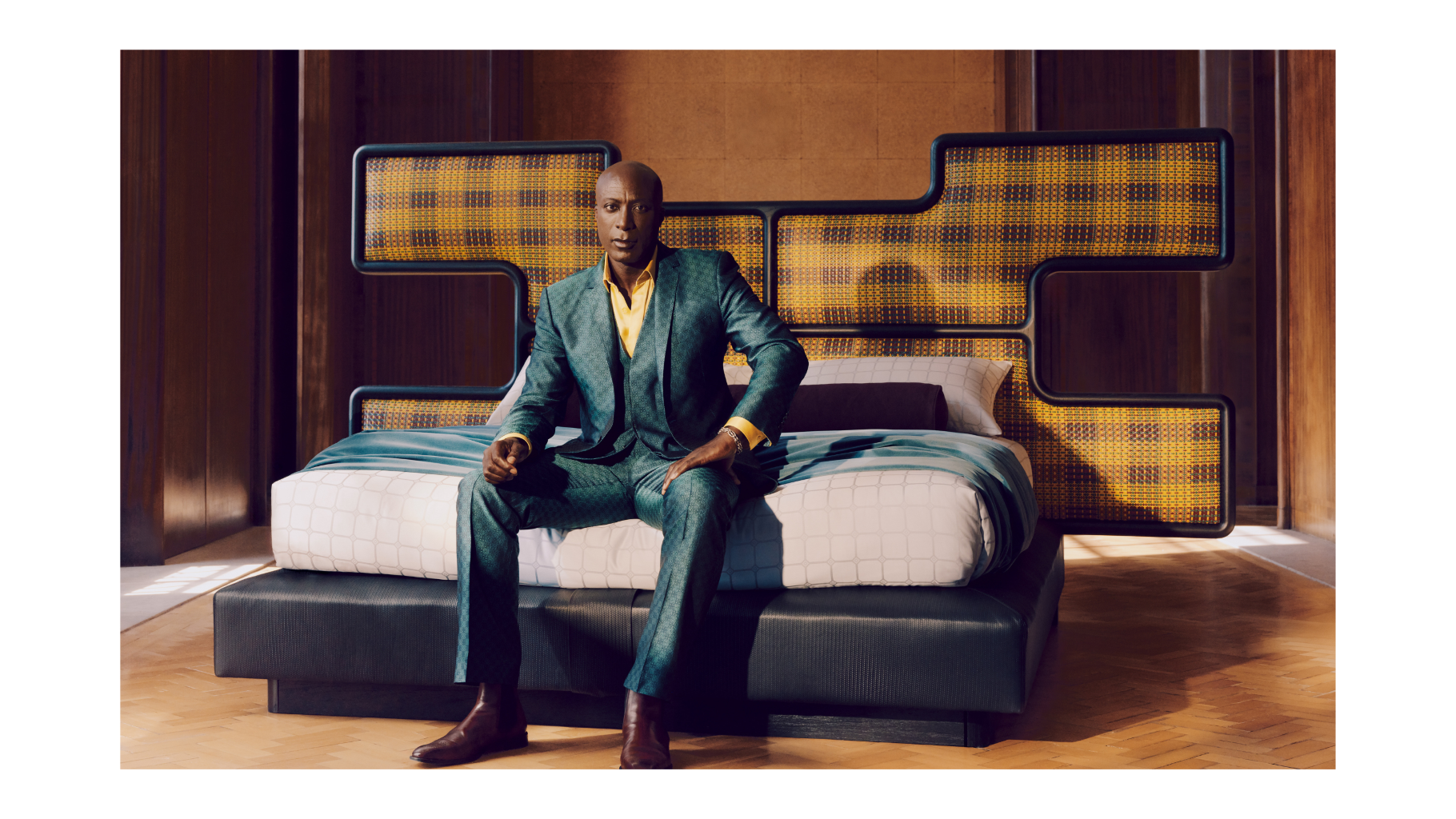 Ozwald Boateng in a suit sitting on a modern bed.