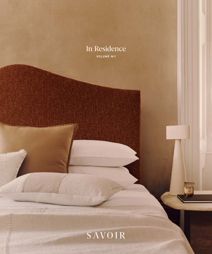 A styled bedroom scene with a bed dressed in white linens, a large rust-brown upholstered headboard, beige and white pillows, a bedside table, a white lamp, and a window. Text on the image reads 'In Residence VOLUME No1 and 'SAVOIR'.