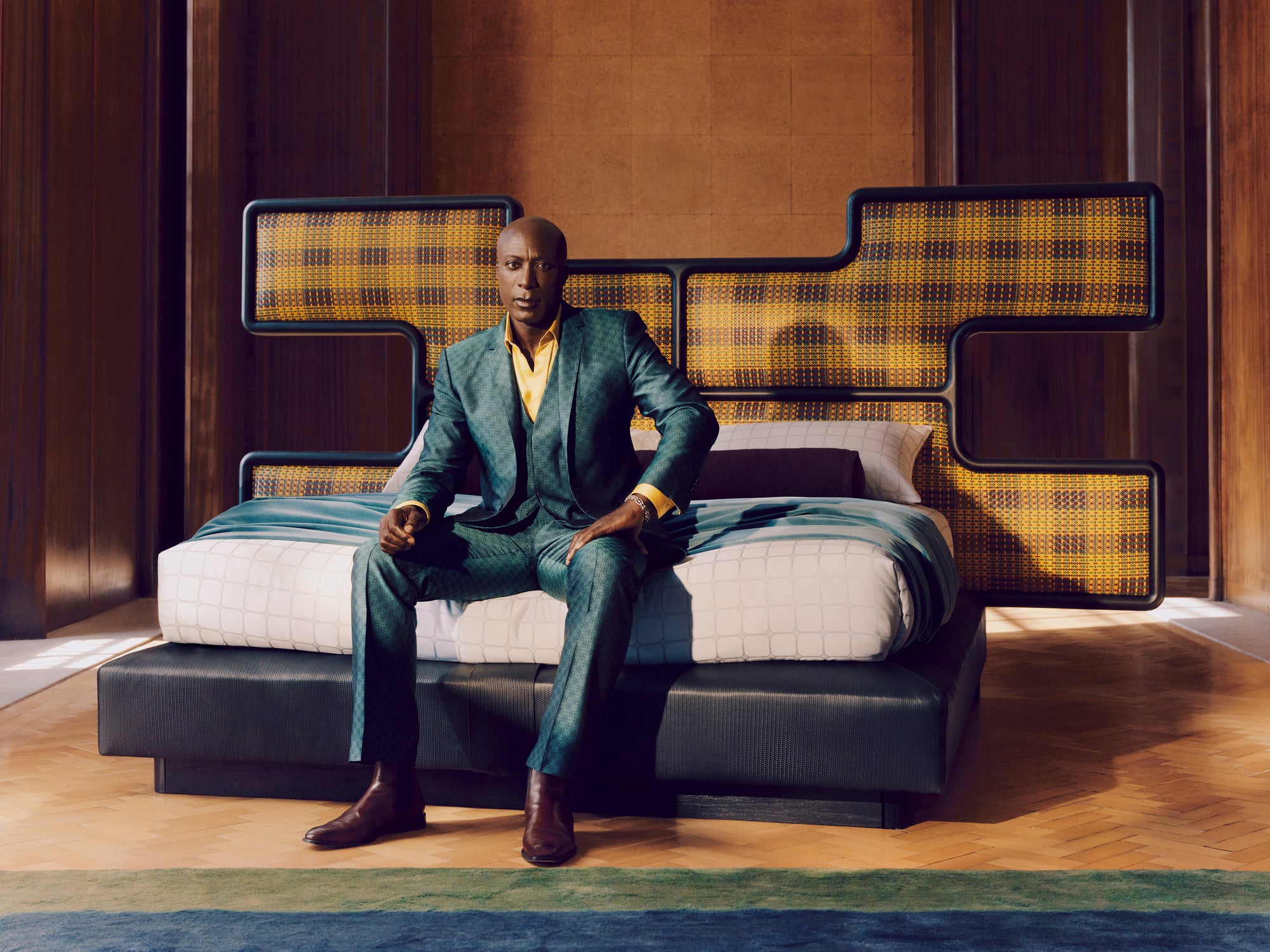 Ozwald Boateng designer in a suit sitting on a modern bed with geometric headboard