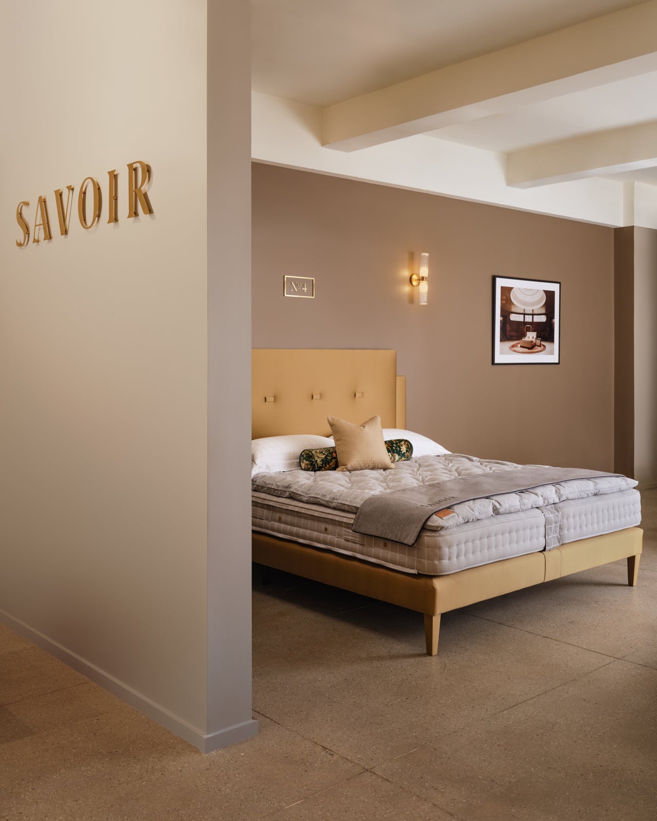 A modern Savoir showroom located in New-York 200 Lex featuring a bed with a light wood frame and tufted headboard, dressed in gray and beige bedding. The showroom has a warm taupe-brown accent wall and a large gold 'SAVOIR' logo on the adjacent cream-colored wall.