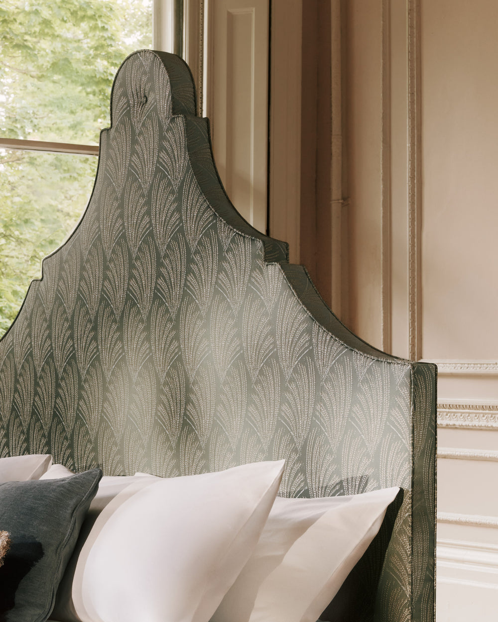 A close-up of a tall upholstered headboard with a scalloped top and a repeating feather-like pattern in muted green and gray tones, shown in a bedroom setting with white pillows and a window behind it.