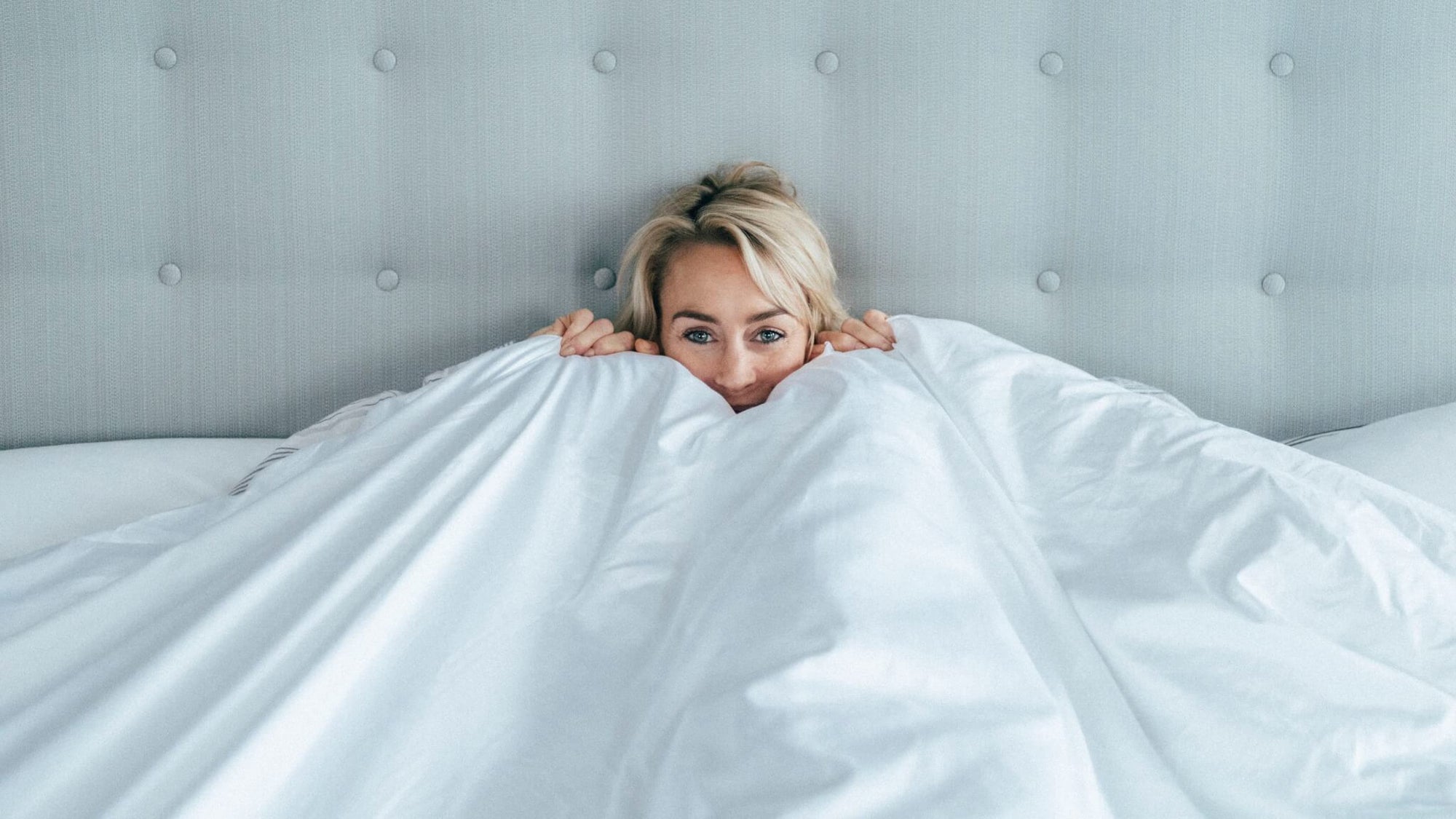 Photo of a blonde woman in a handmade king sized bed holding a duvet up to her face