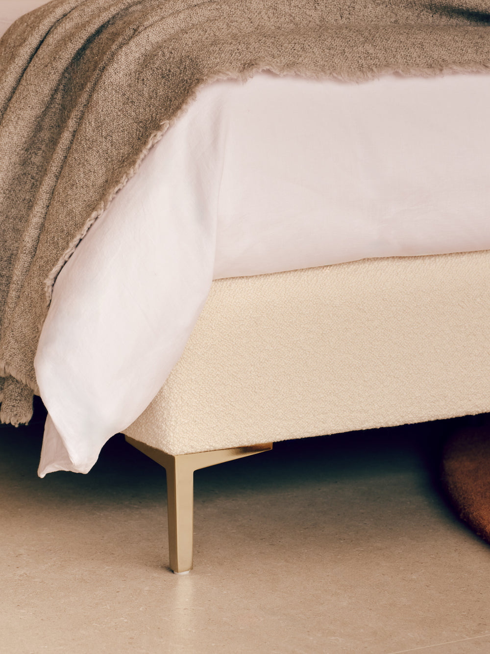 The close-up of the Melrose base bed with a textured cream upholstered base and slim metallic gold legs, dressed in white bedding and a taupe throw blanket.