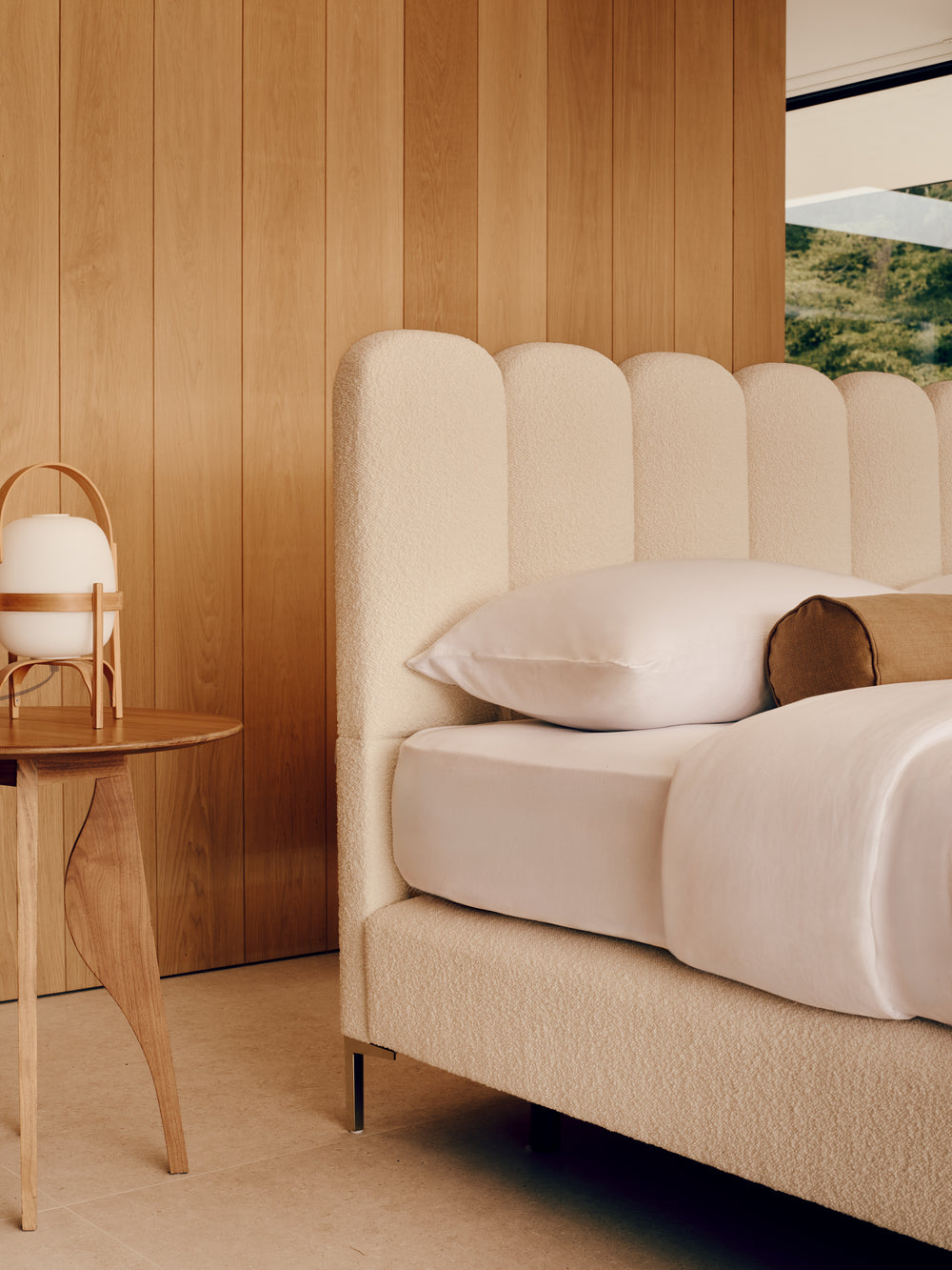 A modern bedroom featuring a textured cream upholstered bed with a scalloped headboard, styled with white linens and a brown accent pillow, set against a warm wood-paneled wall.