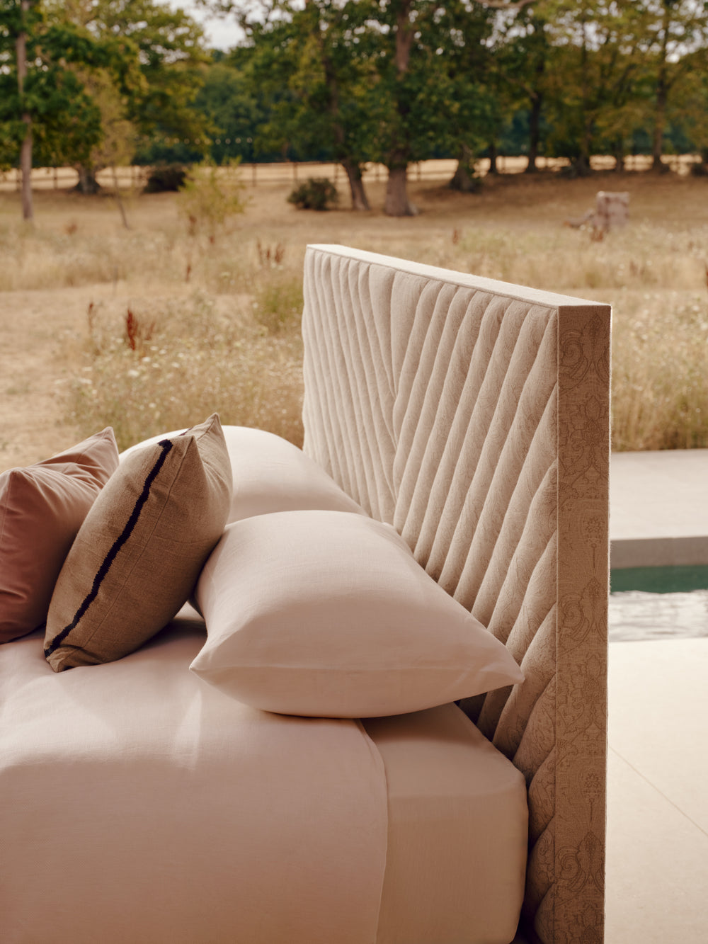 A close-up of the Stella cream headboard with bedding, beige pillows. Set in an outdoor patio area with a blurred grassy landscape and trees in the background.