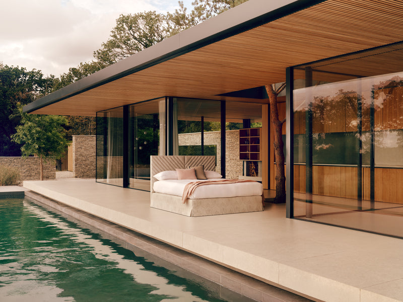 A modern outdoor bedroom setup beside a swimming pool, featuring the Stella bed with white bedding, large floor-to-ceiling glass walls, a wood slatted ceiling overhang, and a stone-and-glass house exterior.