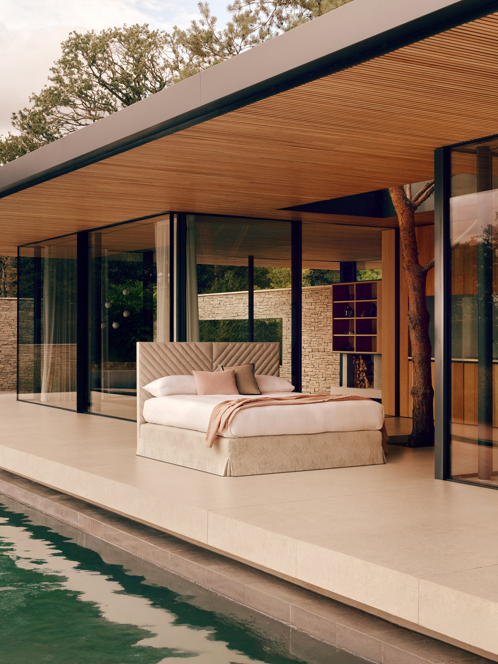 The Savoir Pamela bed with white bedding and a beige upholstered frame sits on a raised patio beside a swimming pool, under a large overhang with wood paneling and floor-to-ceiling glass windows.