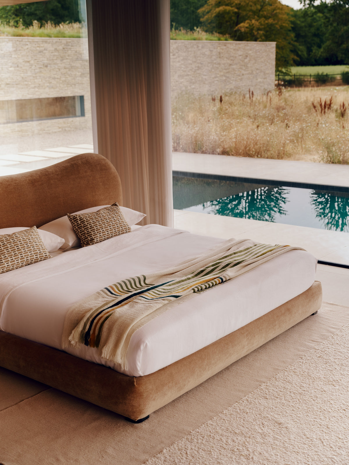 The Teddy bed with a low upholstered frame in warm taupe velvet, dressed in white bedding and a striped throw blanket, set in a minimalist bedroom with a large window overlooking a pool and outdoor landscape.