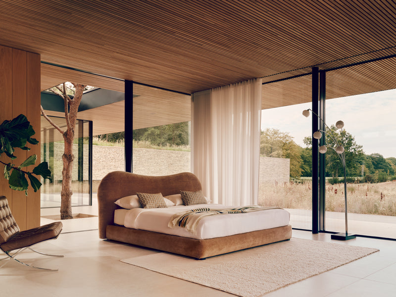 A modern bedroom featuring the curved upholstered Teddy bed in warm brown fabric with white bedding, set in a spacious room with large floor-to-ceiling windows, a wood slat ceiling, and an outdoor view.